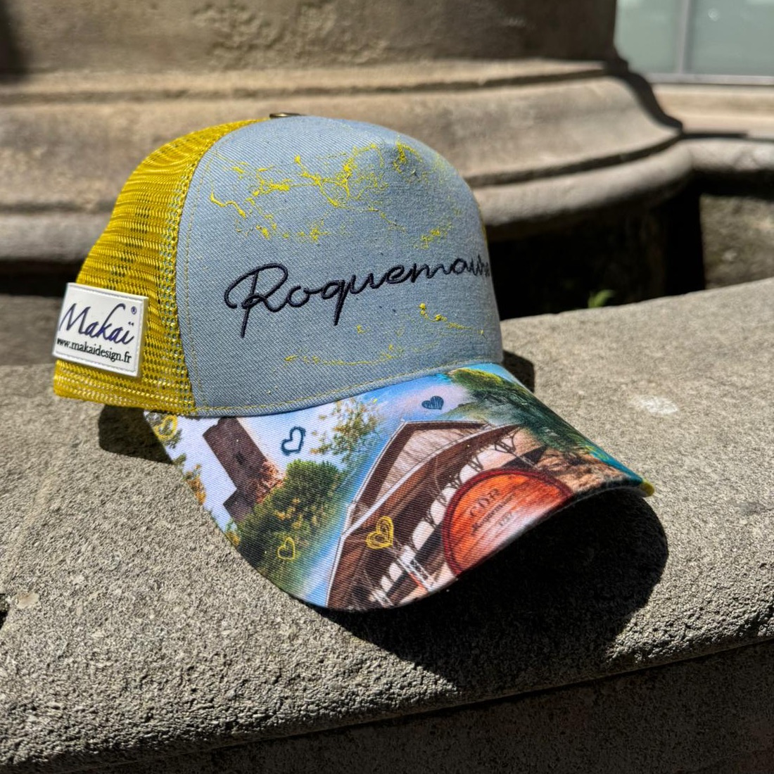 Casquette grise à filet jaune, brodée « Roquemaure », dont la visière illustrée montre la tour carrée, les halles de la Pousterle, un tonneau CDR et des cœurs