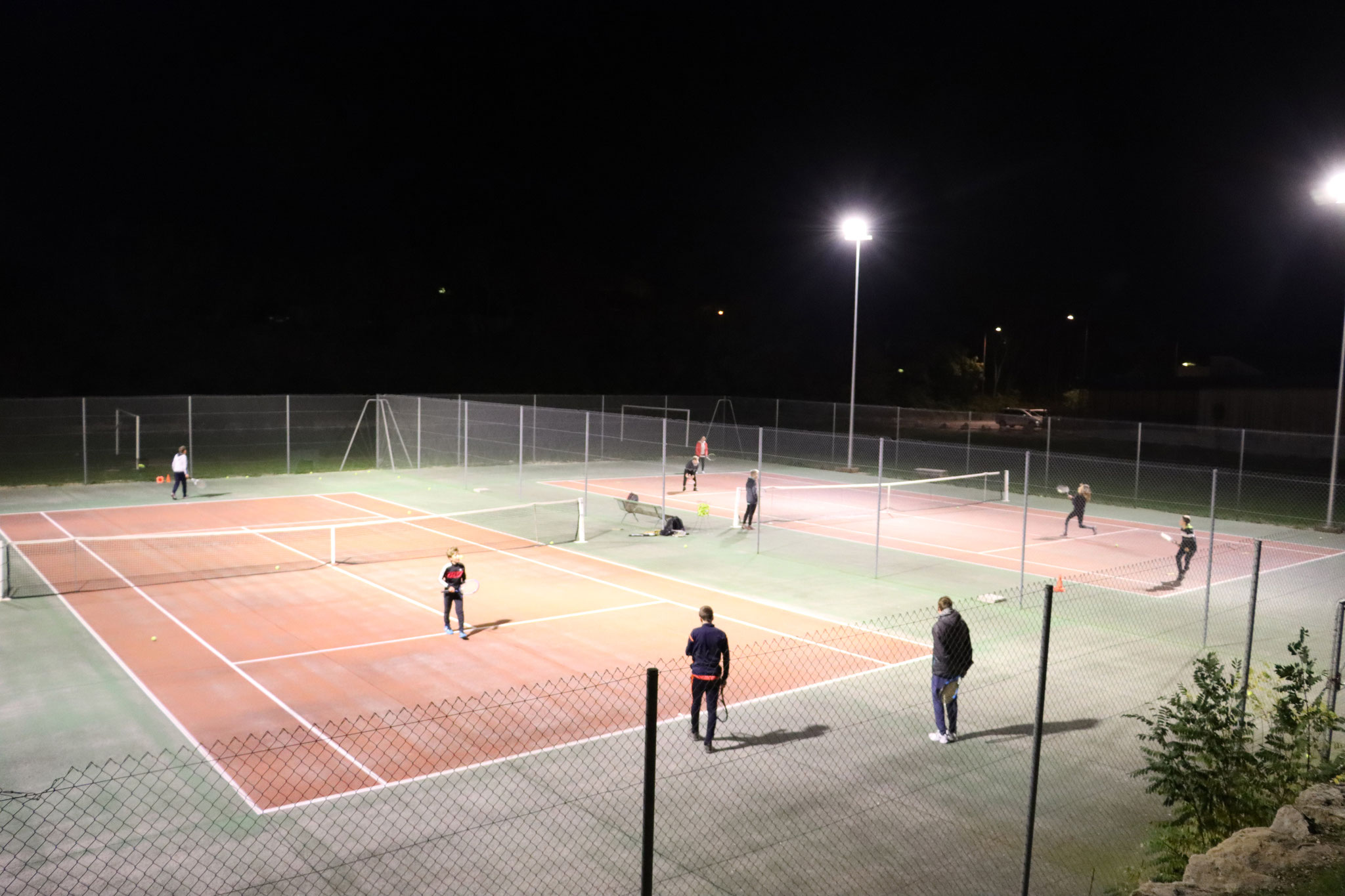 Courts de tennis en résine éclairés par des projecteurs, de nuit, quelques joueurs au loin
