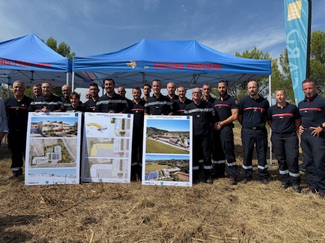 Sapeurs-pompiers en tenue debout sous des barnums, derrière trois panneaux présentant le projet de la future caserne