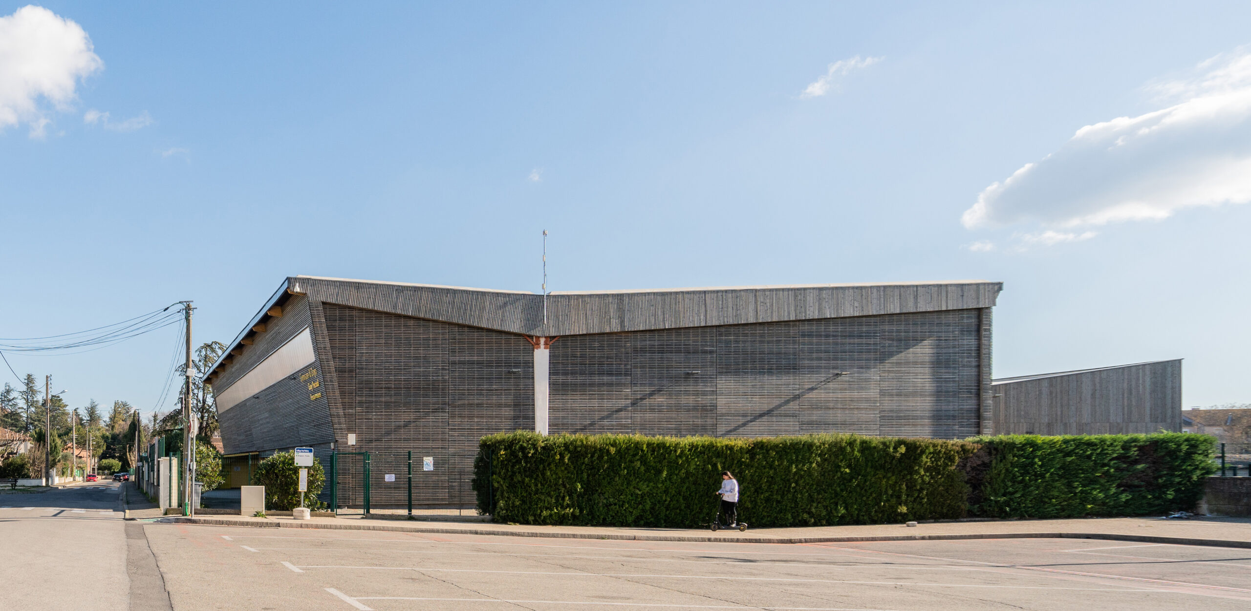 Vue d’ensemble du gymnase en bardage bois depuis la rue, derrière une haie