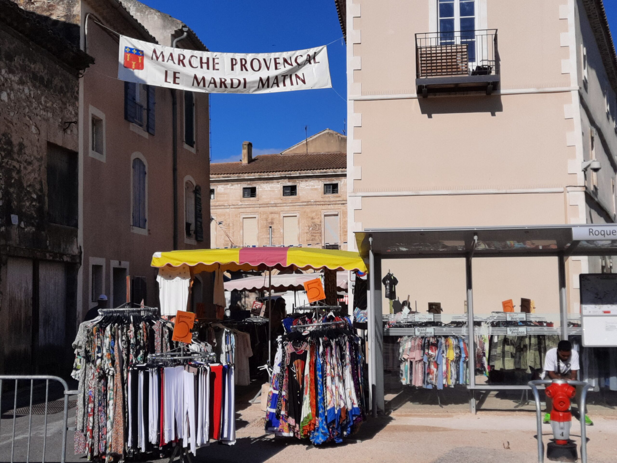 Banderole « Marché provençal le mardi matin » tendue entre deux façades, au-dessus des étals de vêtements