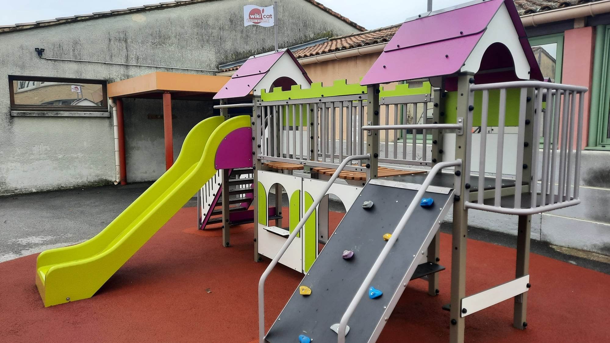 Aire de jeux de l’école maternelle : toboggan vert, plateformes à toits violets et rampe d’escalade sur un sol souple rouge