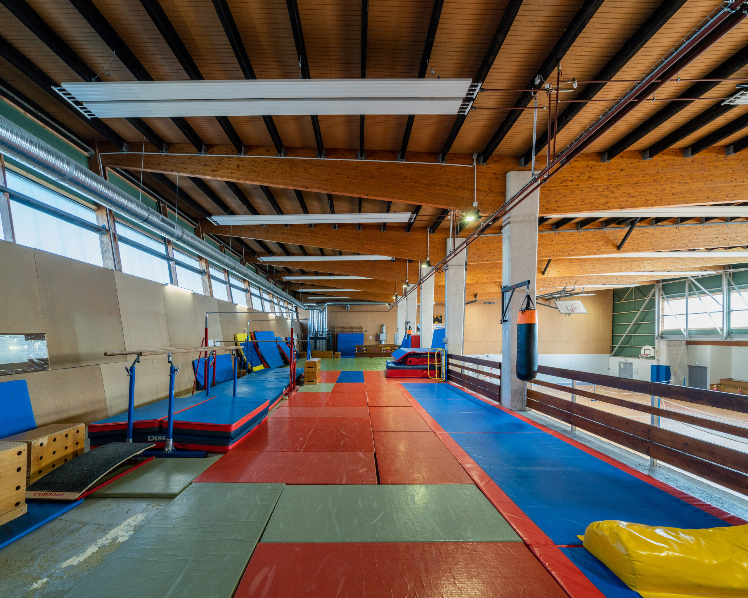 Salle du gymnase sous charpente bois, équipée de tapis de gymnastique et d’agrès
