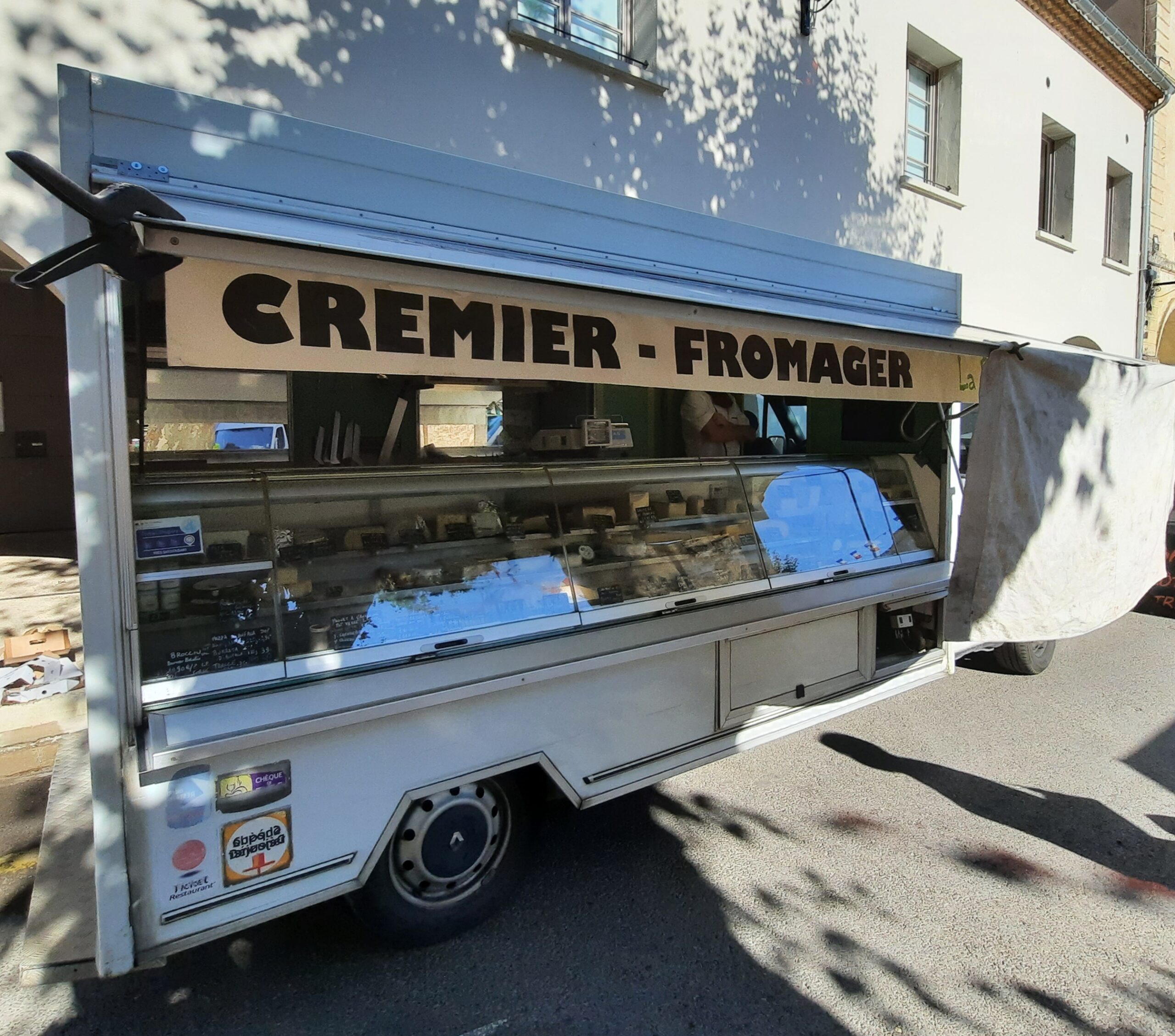 Camion-étal d’un crémier-fromager, vitrine réfrigérée ouverte, garé au soleil devant une façade