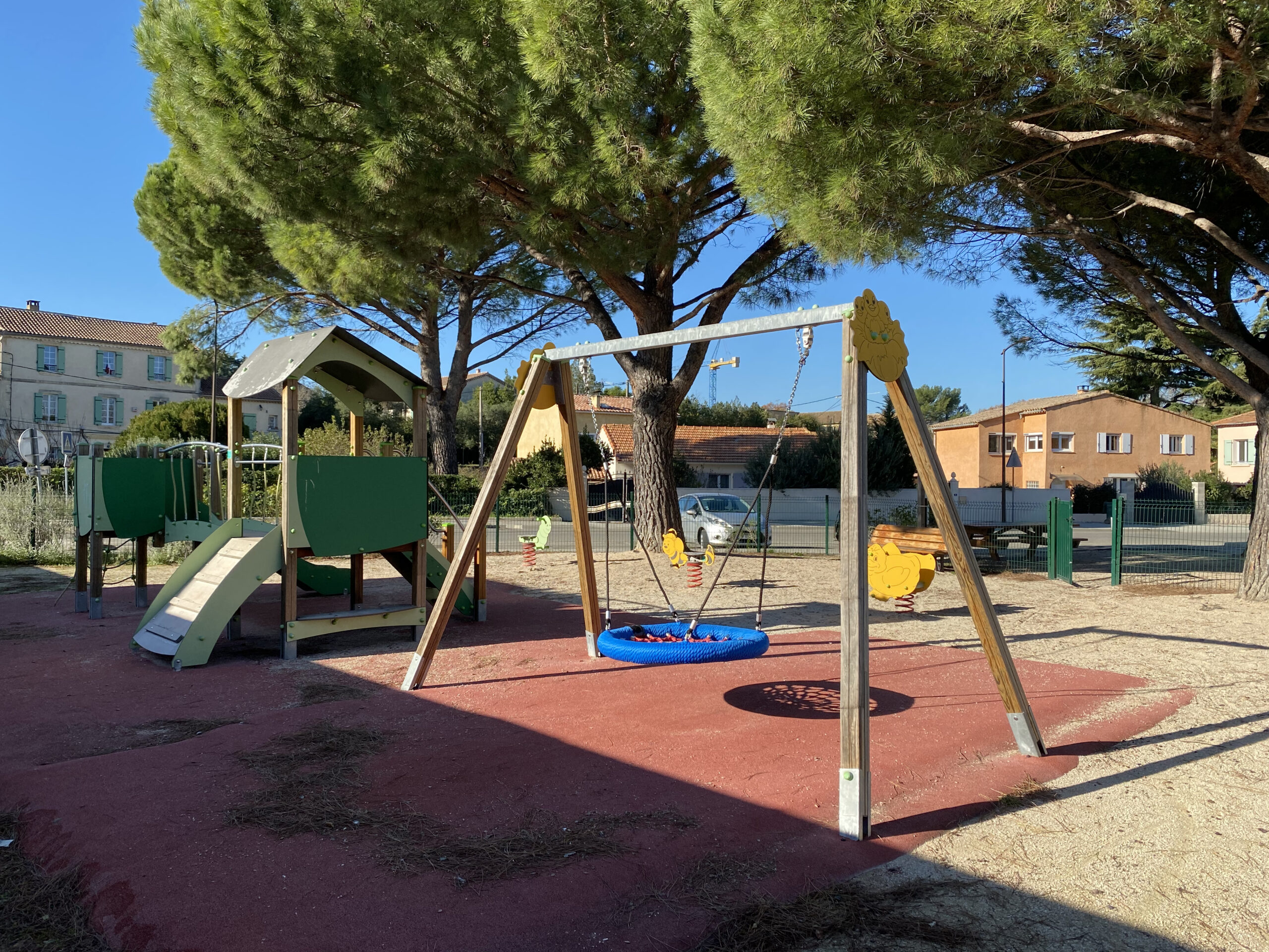 Aire de jeux de la route de Nîmes : balançoire nid d’oiseau et structure à toboggan sur un sol souple rouge, à l’ombre des pins