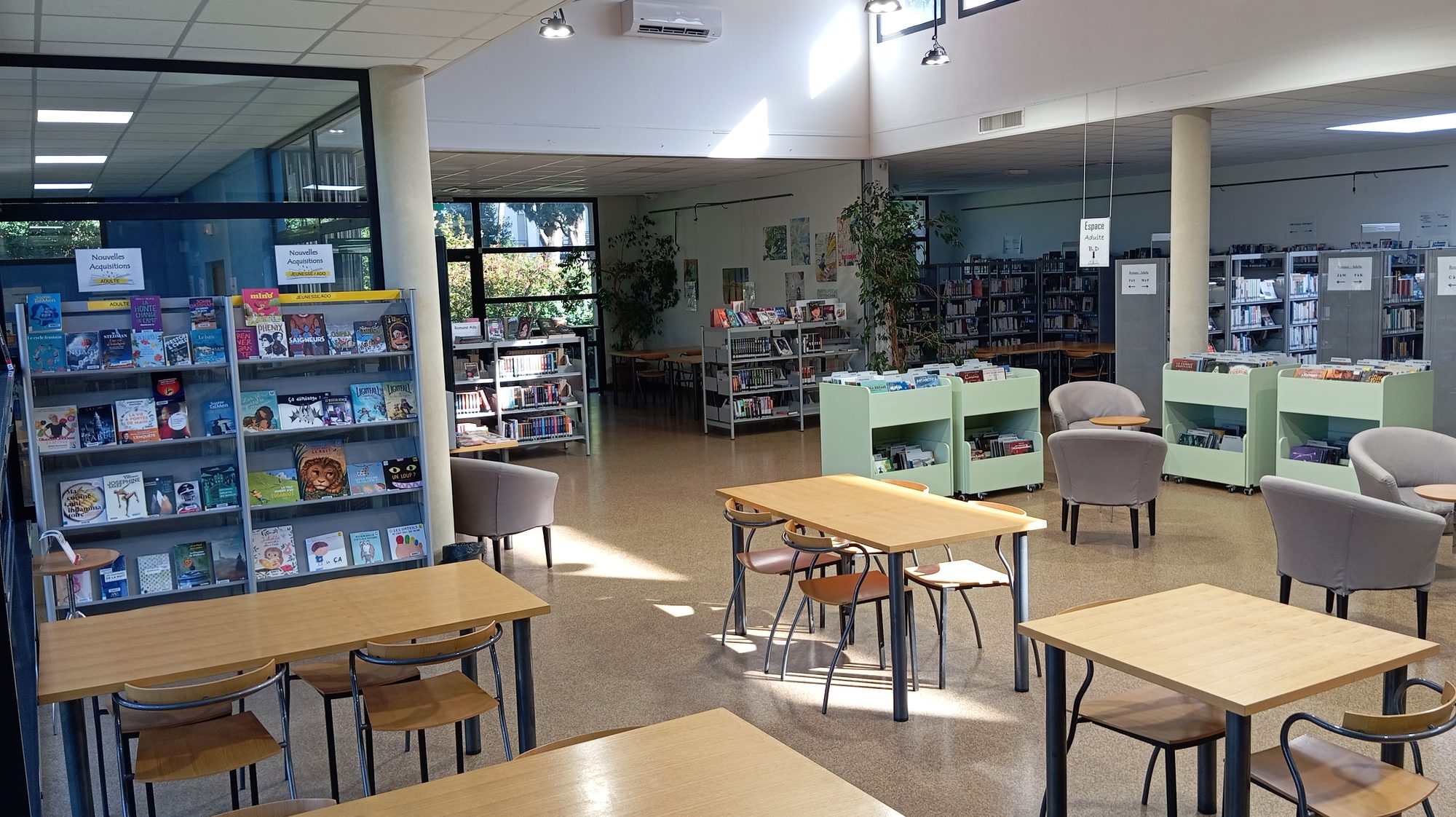 Intérieur de la médiathèque Marc-Alyn : présentoirs de livres, bacs, fauteuils et tables de lecture sous une grande baie vitrée