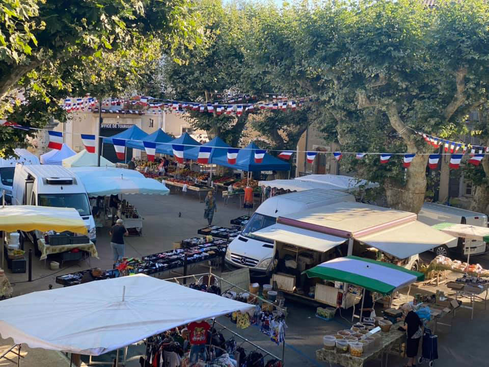 Vue plongeante sur les étals et les camions du marché hebdomadaire, sous les platanes et les guirlandes tricolores