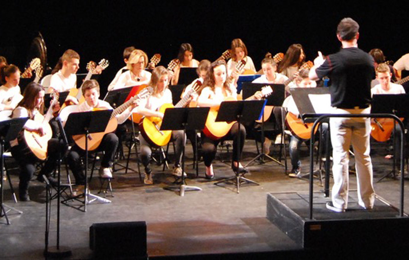 Jeunes guitaristes du conservatoire assis sur scène face à leur chef, de dos, sous les projecteurs