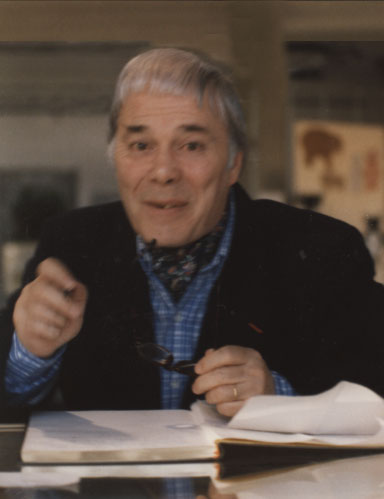 Portrait de Marc Alyn, cheveux blancs, foulard au cou, assis devant un livre ouvert