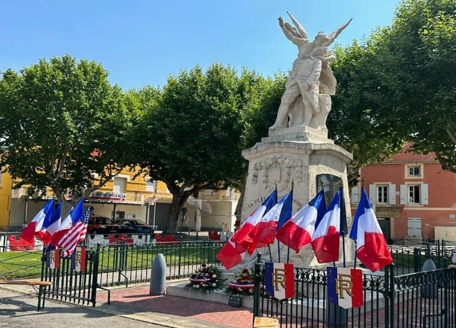 Le monument aux morts pavoisé de drapeaux tricolores à son ancien emplacement, place de la Pousterle, entouré de sa grille, sous les platanes