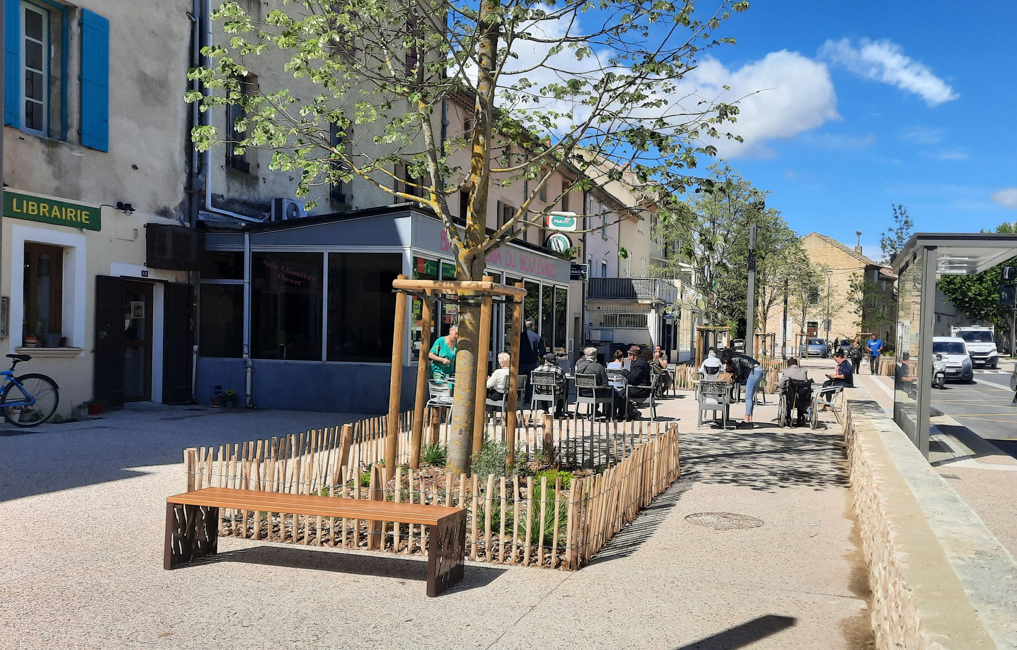 Rue commerçante du centre-ville : librairie, terrasse de café, banc et jeune arbre protégé par une ganivelle