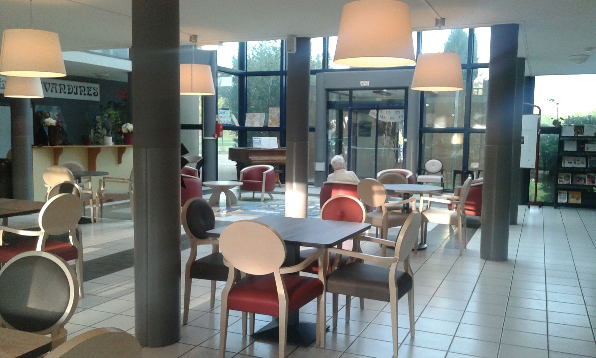 Grand espace commun lumineux de l’EHPAD Les Lavandines, tables, chaises rouges et suspensions, baies vitrées au fond