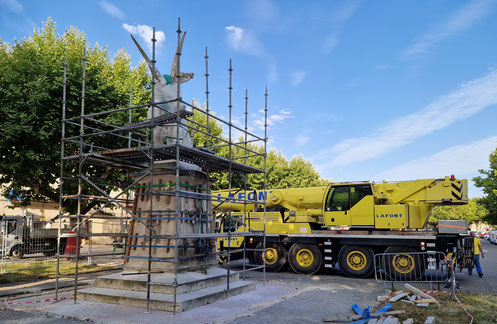 Le monument aux morts sous un échafaudage, à côté d’une grue jaune, place de la Pousterle