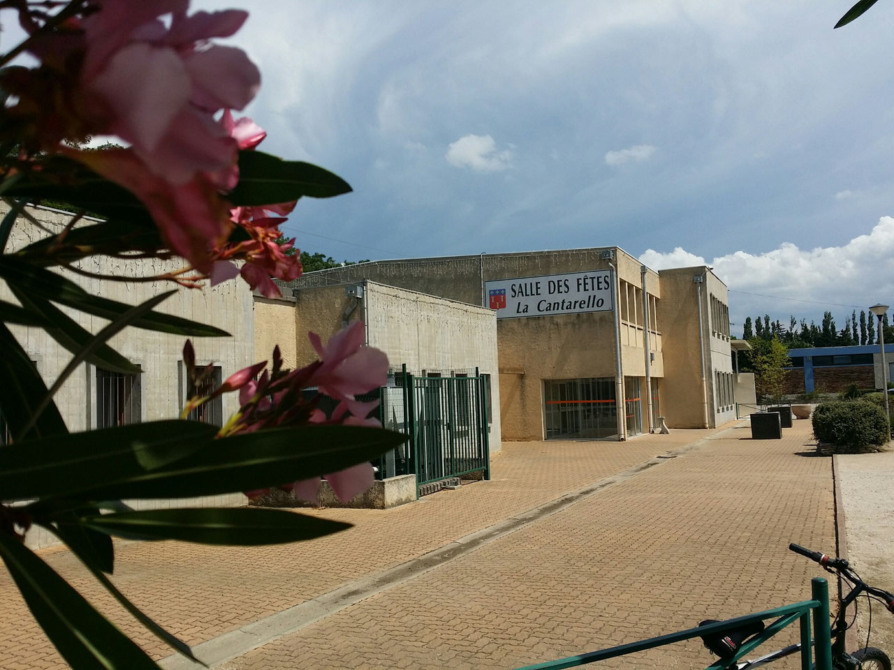 Bâtiment de La Cantarello avec son enseigne « Salle des fêtes La Cantarello », parvis pavé et laurier-rose en fleur au premier plan
