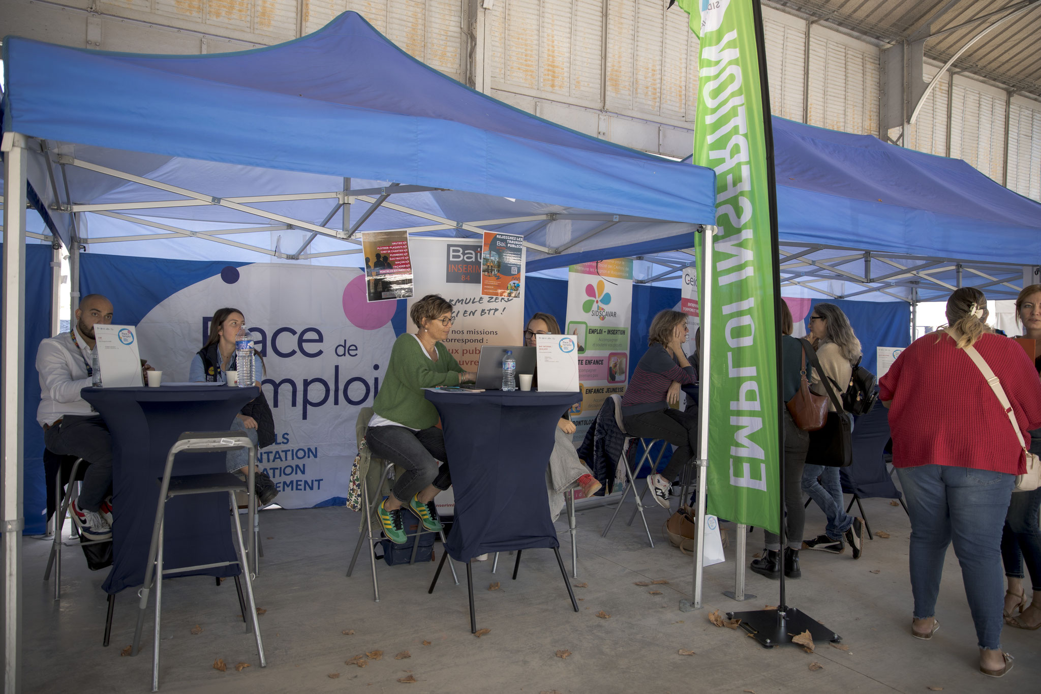 Conseillers attablés sous des barnums bleus, kakémonos « Place de l’emploi » et « Emploi », visiteurs à leurs côtés