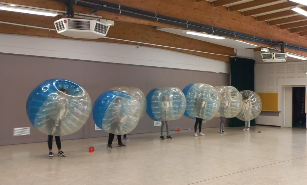 Six jeunes dans des bulles gonflables transparentes, alignés dans une grande salle