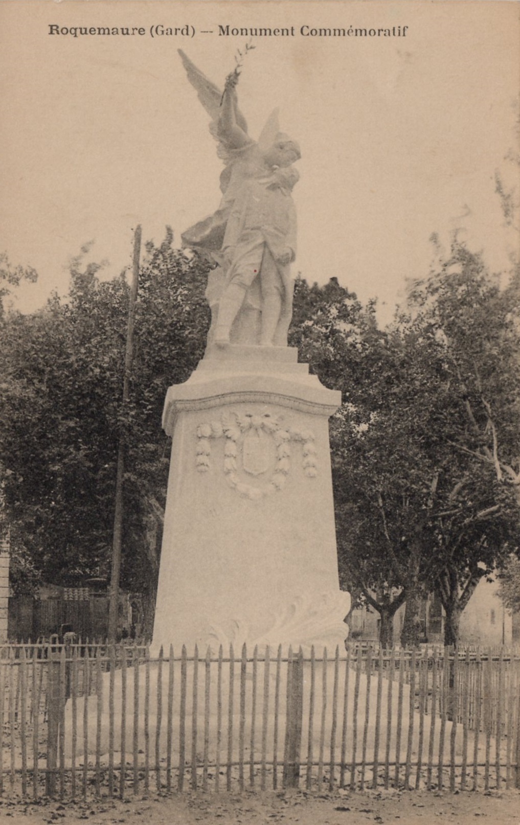 Carte postale ancienne « Roquemaure (Gard), Monument commémoratif » : la statue ailée sur son haut socle aux armoiries, derrière une palissade, devant les arbres de la place