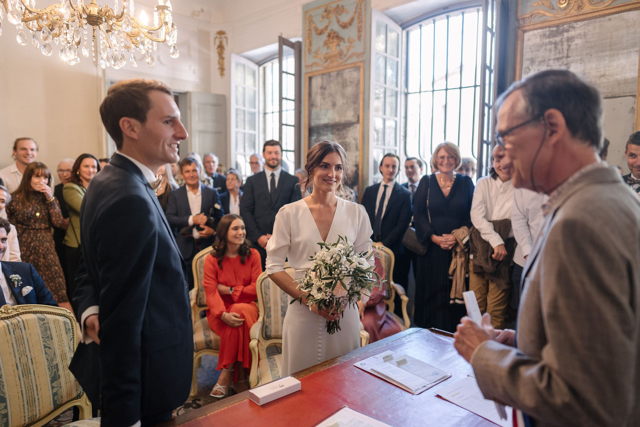 Les futurs époux face à l’officier d’état civil dans la salle des mariages, sous les lustres, devant leurs invités